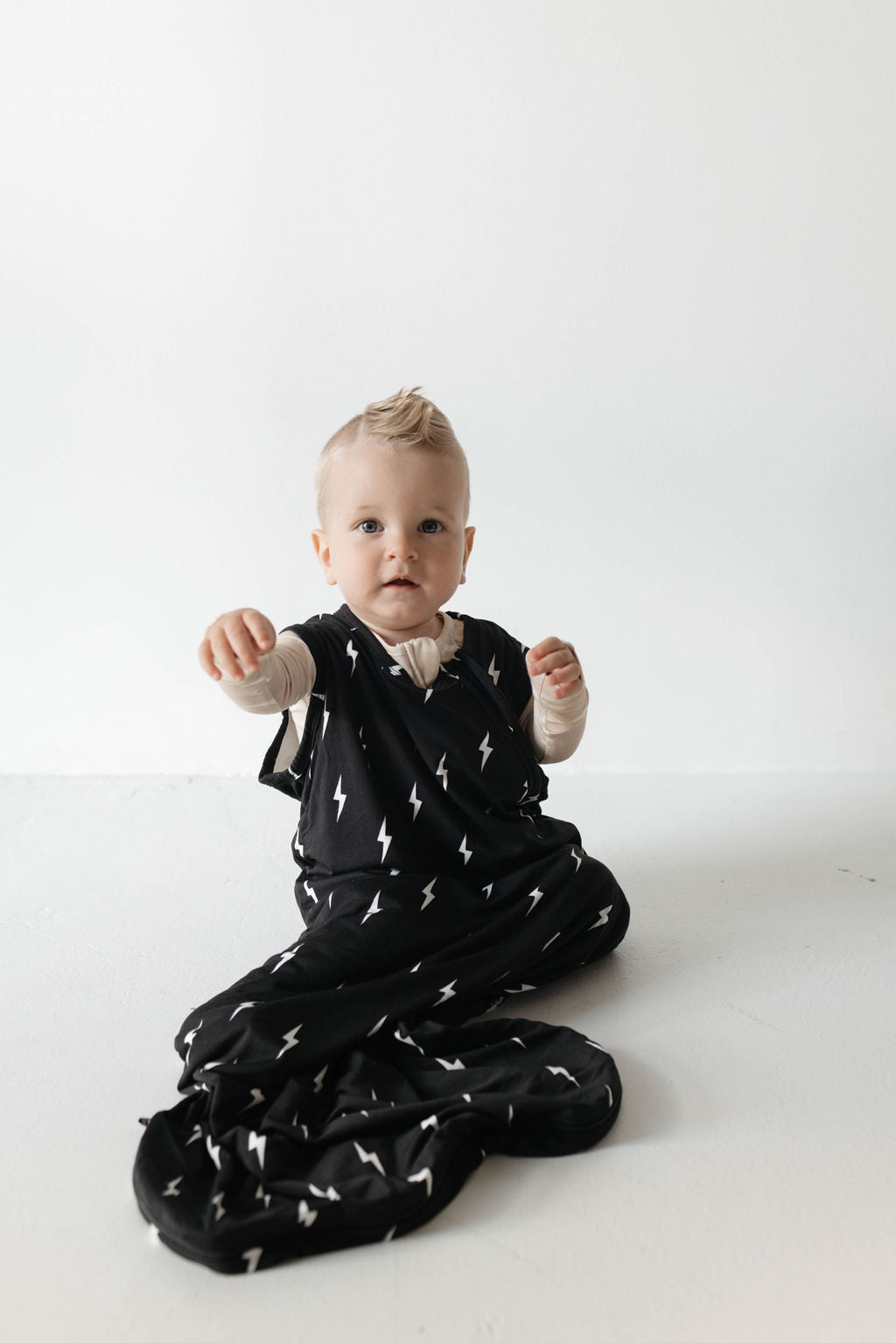 A baby with light hair is sitting on the floor, wearing a black onesie adorned with white lightning bolt patterns and has light-colored sleeves. The baby is reaching out with both hands while wrapped in the luxurious Bamboo Sleep Sack | Midnight Lightning Bolt by forever french baby. The background is plain and white.