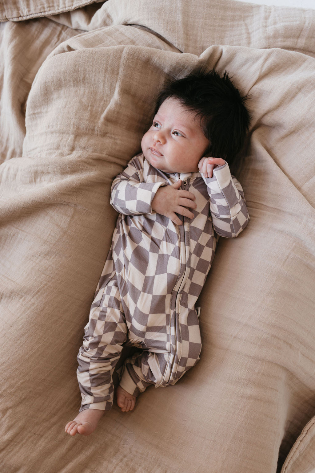 A baby with dark hair lies comfortably on a beige, textured blanket wearing Bamboo Zip Pajamas in Smokey Wave from forever french baby, looking off to the side with a relaxed expression.