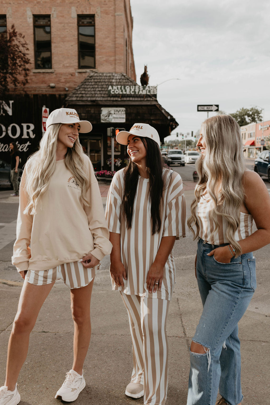 Three women stand on a sidewalk, smiling and engaging with each other. They are wearing casual clothing: two in matching striped outfits and one in jeans with a striped top. All three wear the Kalispell Trucker Hat by forever french baby, discussing product availability, while the building in the background has a vintage storefront appearance.