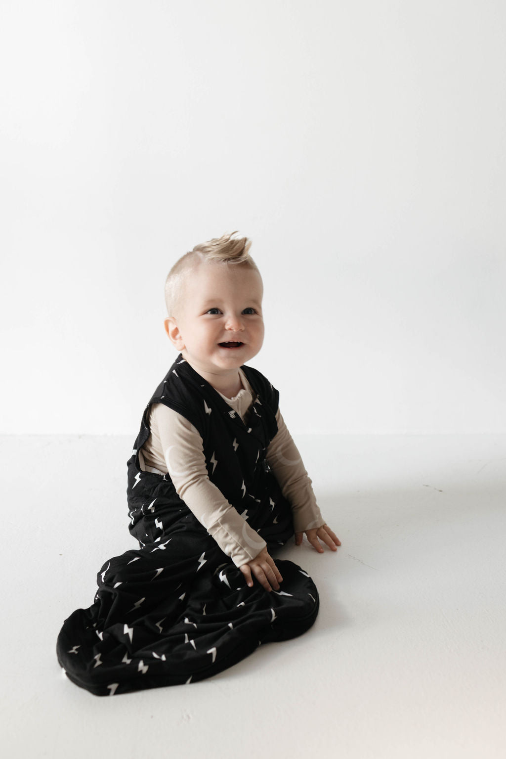 A small child sits on the floor, wearing the Bamboo Sleep Sack in Midnight Lightning Bolt from forever french baby and a beige long-sleeve shirt underneath. The child has short blonde hair styled in a small fauxhawk and is smiling while looking slightly off to the side. They look ready for luxurious baby sleep in their stylish wearable blanket.