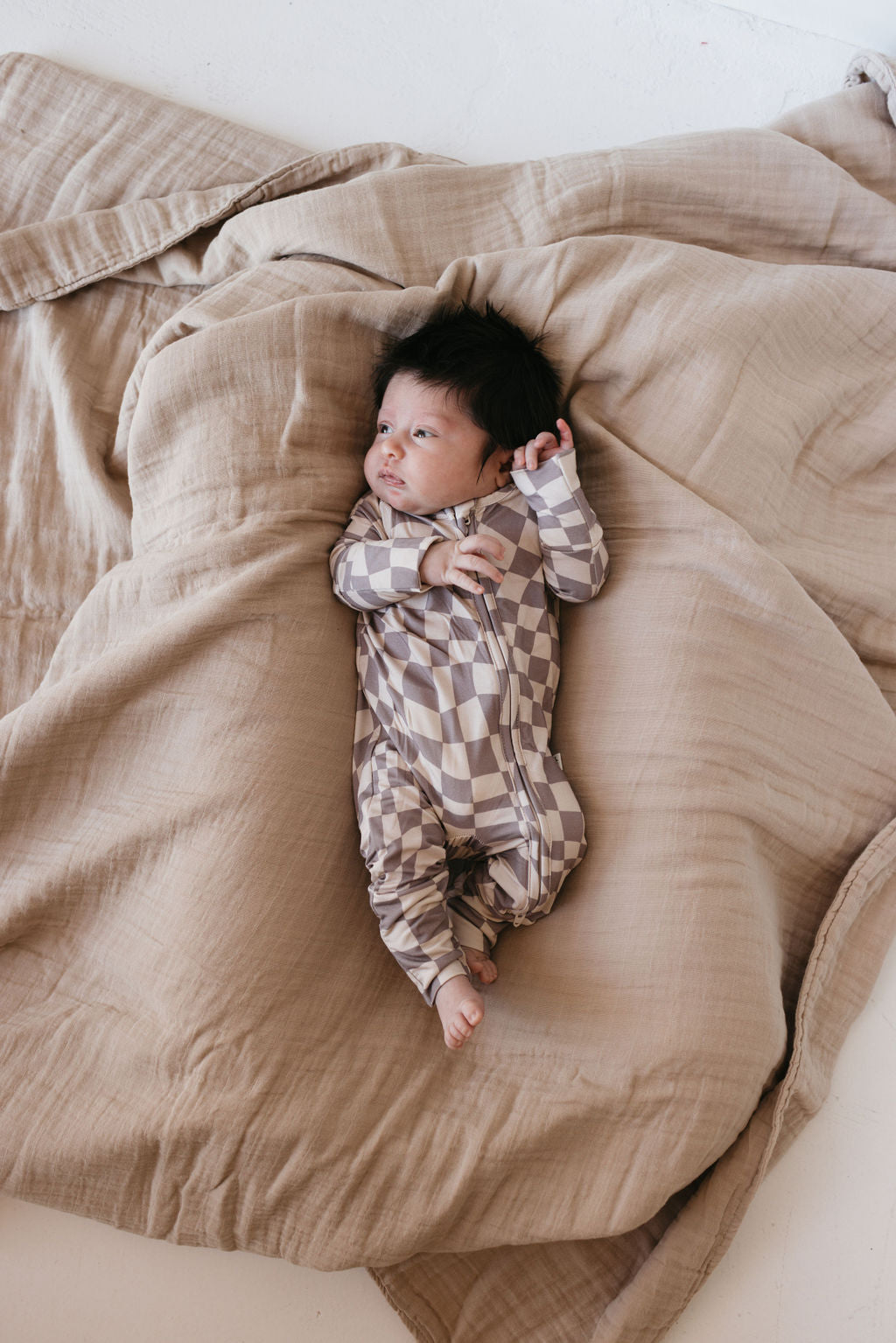 A baby wearing forever french baby's Bamboo Zip Pajamas in the Smokey Wave design lies on a soft, beige blanket. The baby has dark hair and looks up with its right hand raised near the head, creating a cozy atmosphere enhanced by the breathable baby wear, which ensures comfort with its soft fabric and gentle colors.