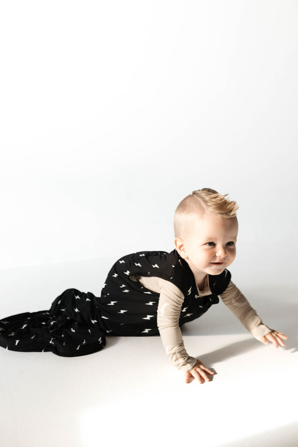 A baby with short blonde hair smiles while crawling on the floor. The baby is wearing a luxurious Bamboo Sleep Sack in Midnight Lightning Bolt from forever french baby, paired with a beige long-sleeve shirt. The background is white and well-lit.