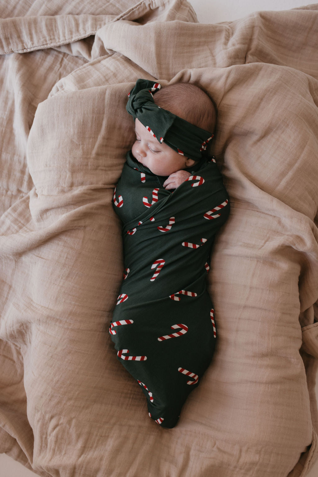 A baby is peacefully sleeping, wrapped in the hypo-allergenic Bamboo Swaddle from forever french baby's Candy Cane Lane collection, featuring a dark green design with candy cane patterns. The bamboo fabric ensures comfort as the baby wears a matching headband against a soft, beige background.