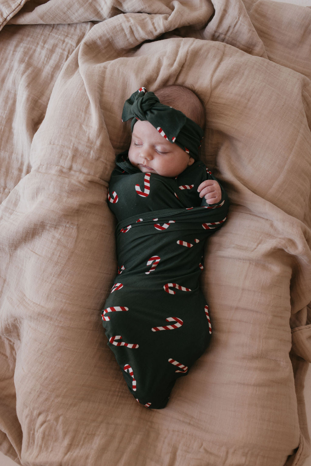 A baby is peacefully sleeping wrapped in the Bamboo Swaddle from the Candy Cane Lane collection by forever french baby, featuring a green fabric adorned with candy cane patterns. They rest on a beige comforter, while the breathable material and matching headband provide added comfort and tranquility.