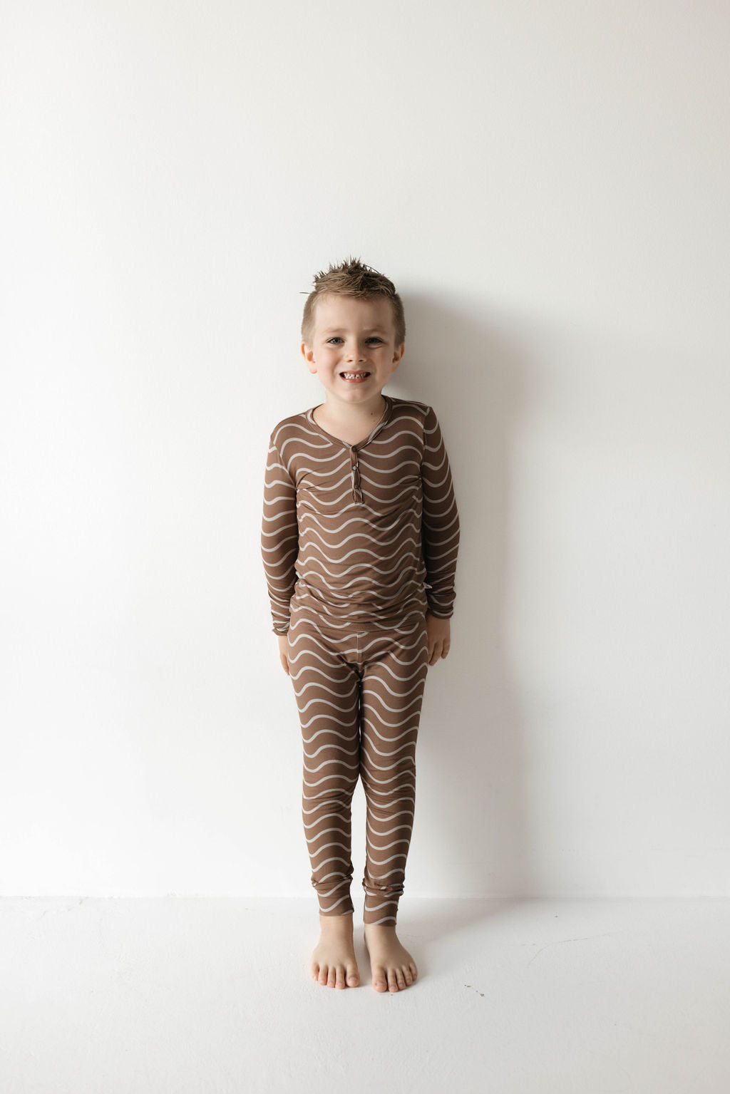 A young child with short brown hair stands against a plain white background, smiling and barefoot, wearing matching long-sleeve forever french baby Bamboo Two Piece Pajamas in the Static Waves pattern.