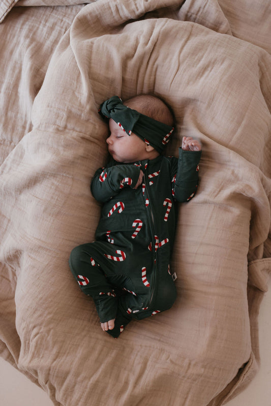 A baby sleeps peacefully on a beige blanket, dressed in Bamboo Zip Pajamas from forever french baby, featuring charming candy cane patterns and accompanied by a matching headband. The breathable attire adds a cozy and festive touch to the scene.