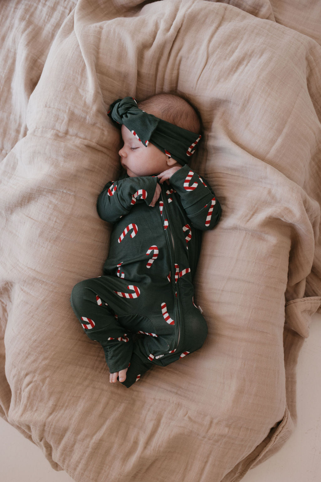 A baby rests peacefully on beige blankets, wearing forever french baby's Bamboo Zip Pajamas in dark green with a Candy Cane Lane pattern. A matching headband adorns the baby's head as they lie serenely on their side.