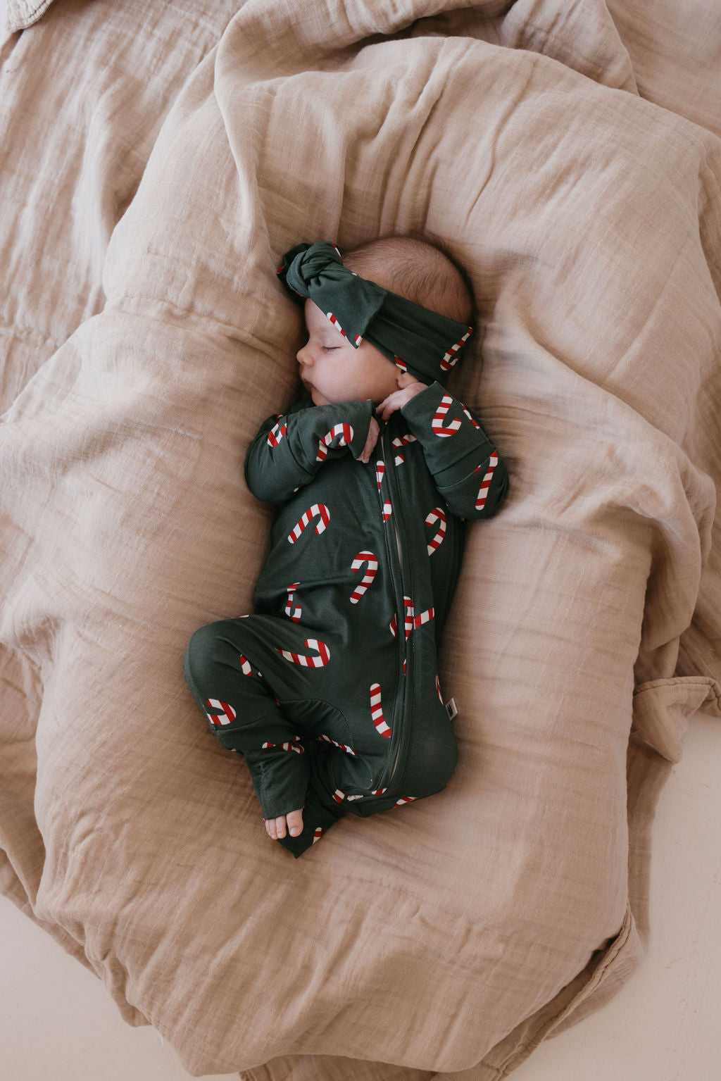 A sleeping baby is comfortably wrapped in beige sheets, wearing forever french baby's Bamboo Zip Pajamas in the Candy Cane Lane design. The baby sports a matching headband and is peacefully resting on their side.