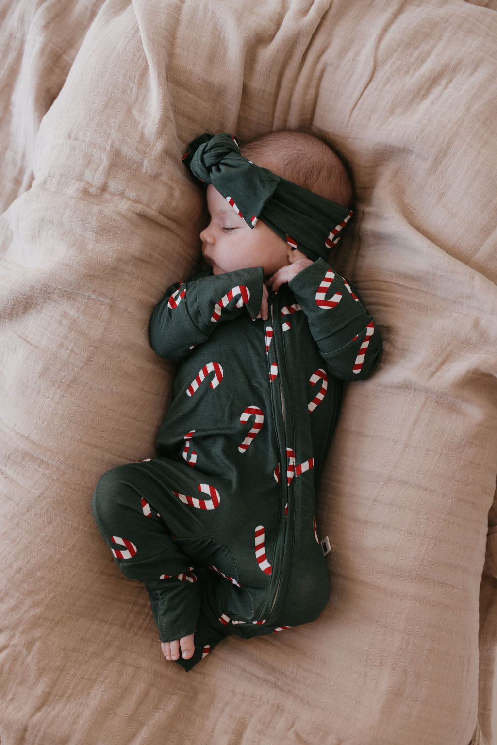 A sleeping baby is dressed in a dark green onesie featuring candy cane patterns, crafted from soft bamboo fabric, and complemented by an adjustable Bamboo Head Wrap in the Candy Cane Lane design. They are comfortably resting on a snug, light brown blanket, encapsulating the comfort and style of forever french baby.