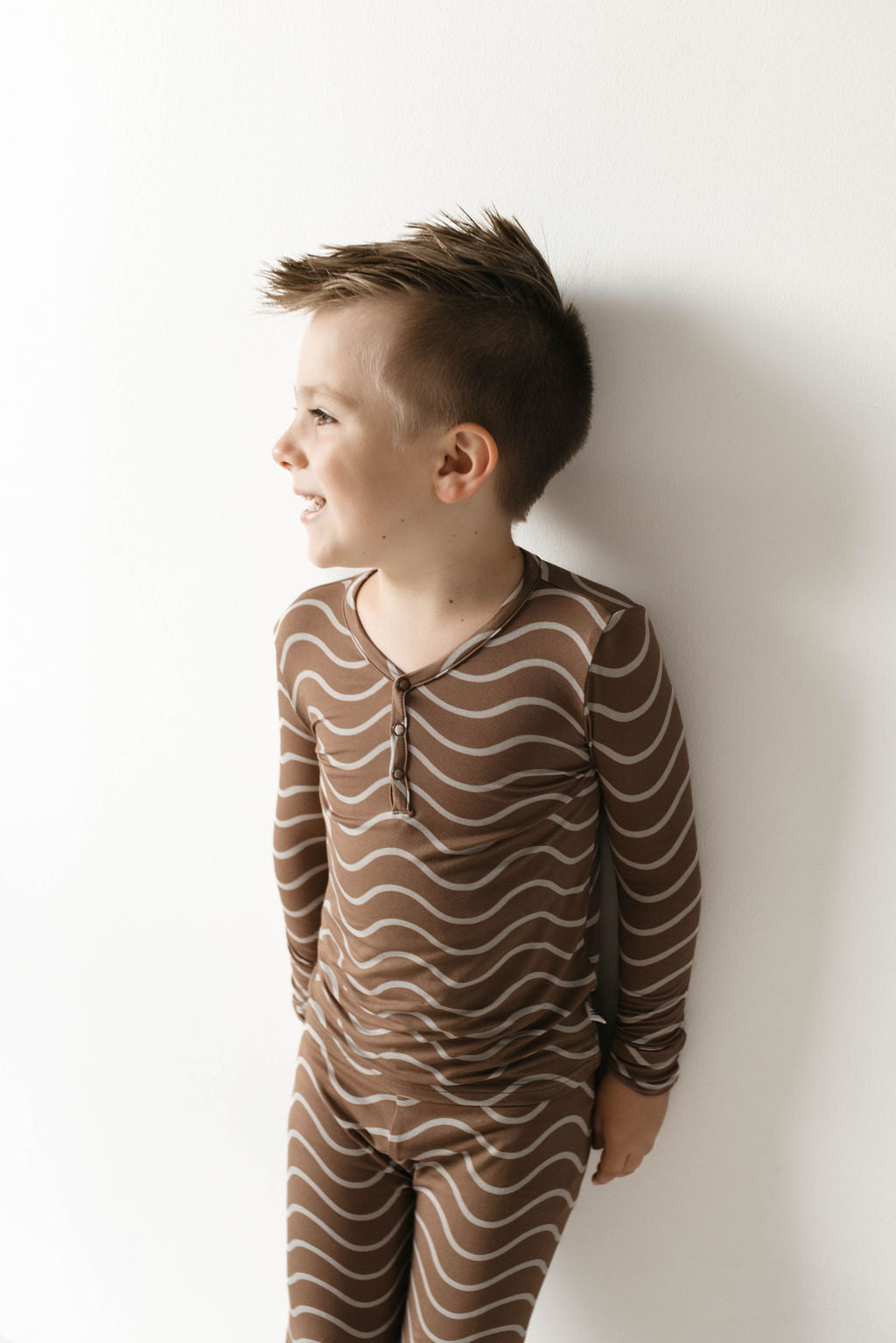 A young boy stands against a white wall, looking to the side and smiling. His short, styled hair complements his outfit: breathable Bamboo Two Piece Pajamas from forever french baby. The matching long-sleeve shirt and pants feature a brown and white wavy stripe pattern named Static Waves. With his hands casually tucked in his pockets, he exudes comfort and style.