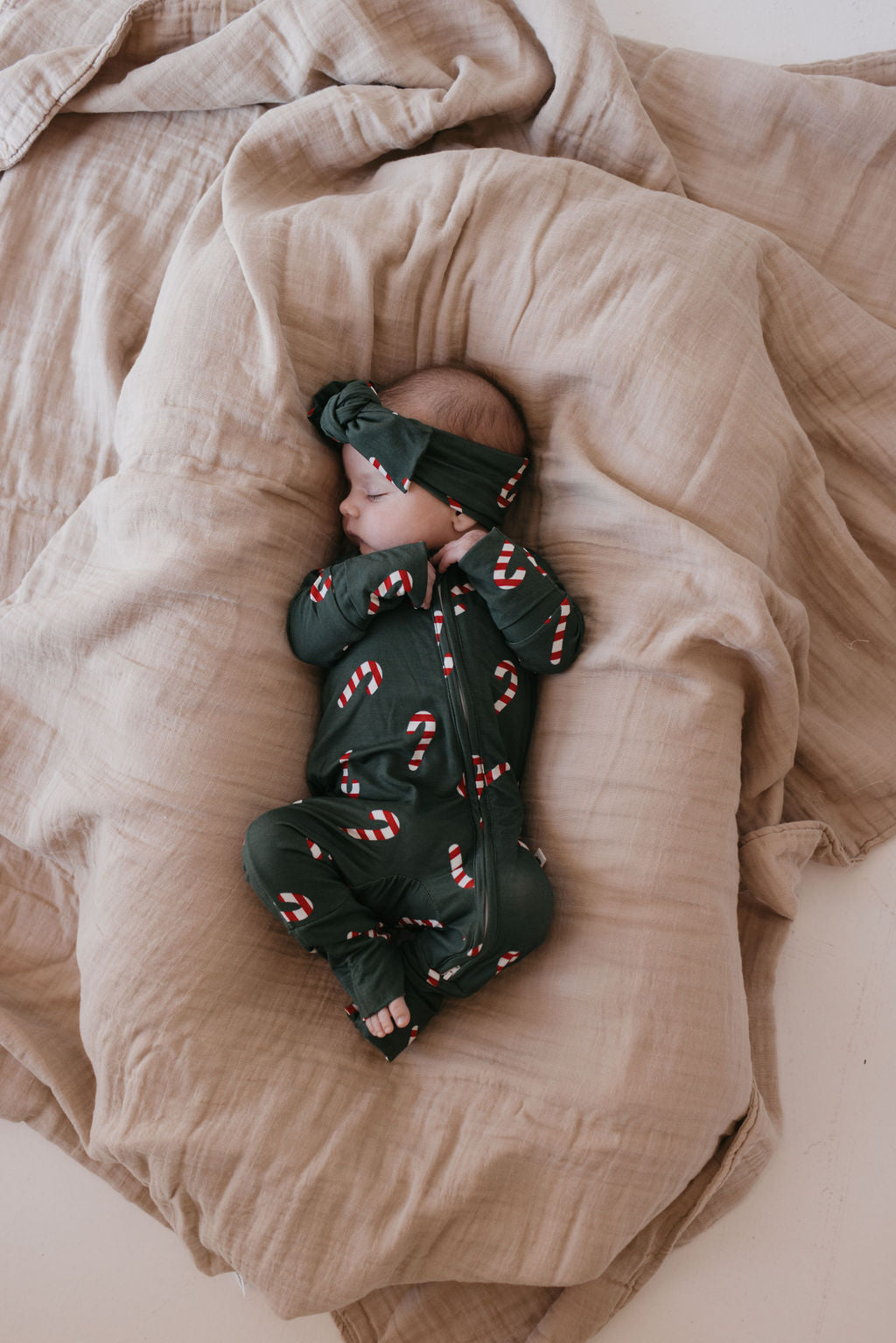 A baby peacefully sleeps on beige blankets, dressed in a dark green onesie with a candy cane pattern and the matching Bamboo Head Wrap from Forever French Baby's Candy Cane Lane collection.