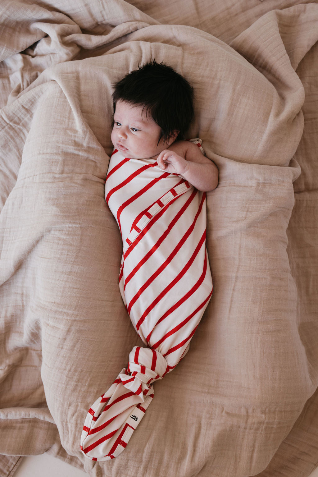 A newborn baby wrapped in a Bamboo Swaddle | the Claus by forever french baby lies on soft, beige bedding. The breathable fabric gently cradles the baby with care, as their dark hair frames a curious gaze upward, one hand near their face.