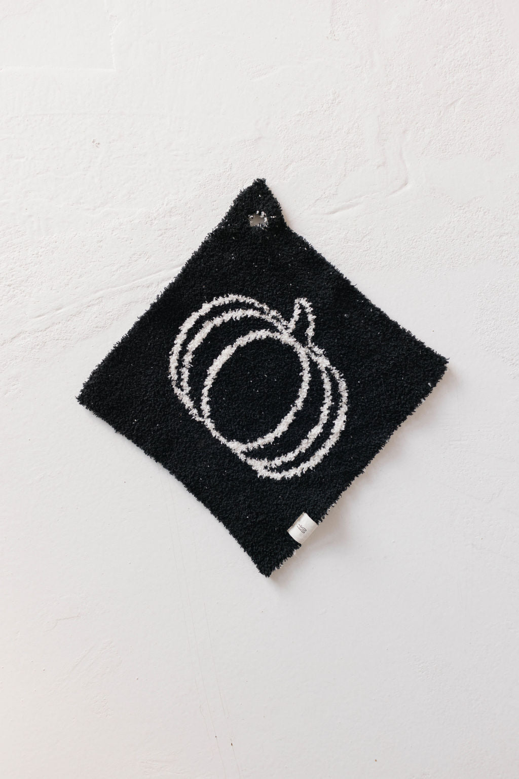 Introducing the **Lovey | Midnight Pumpkin** by **forever French baby**: A black square potholder adorned with a white outline drawing of a pumpkin at its center. This plush accessory is crafted from soft microfiber feather yarn, complete with a small hanging loop at the top, and showcased against a white textured wall.