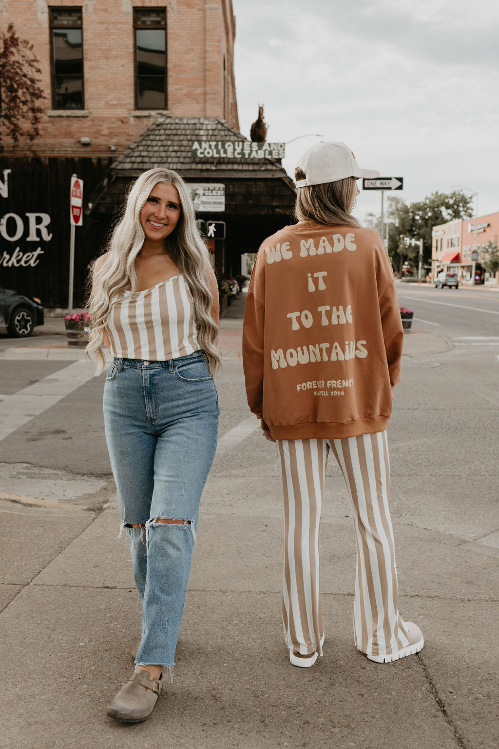 Two people stand on a sidewalk in a small town. One wears jeans and a striped top, smiling at the camera. The other sports striped pants made of 95% cotton and elastane, a hat, and an Adult Sweatshirt from forever French baby with the text "Made It to The Mountains." The buildings in the background include a market and a coffee shop.