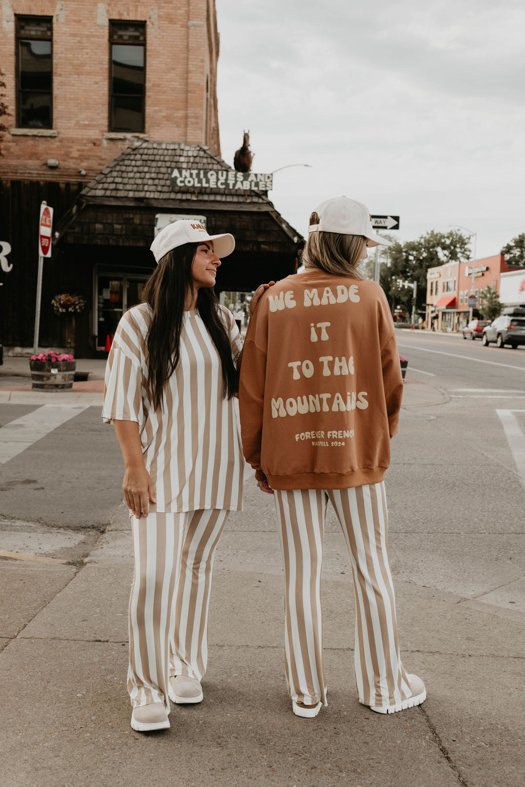 Two individuals stand at a street corner, both dressed in matching striped outfits made from 95% cotton and elastane, coupled with white caps. One faces forward while the other, back to the camera, sports the "Adult Sweatshirt | Made It to The Mountains" by forever French baby. Brick buildings and a sign sit in the background.