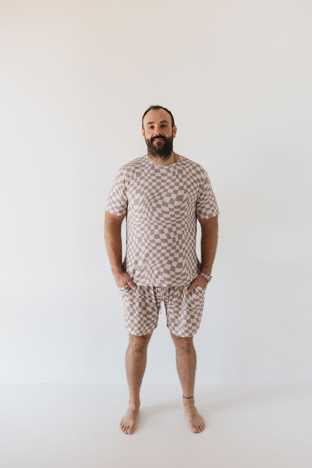 A bearded man stands barefoot against a plain white background, wearing the Men's Bamboo Short Pajamas in Smokey Wave from forever french baby. His hands rest in his pockets, and he sports a relaxed expression, savoring the hypo-allergenic comfort of the breathable bamboo fabric.