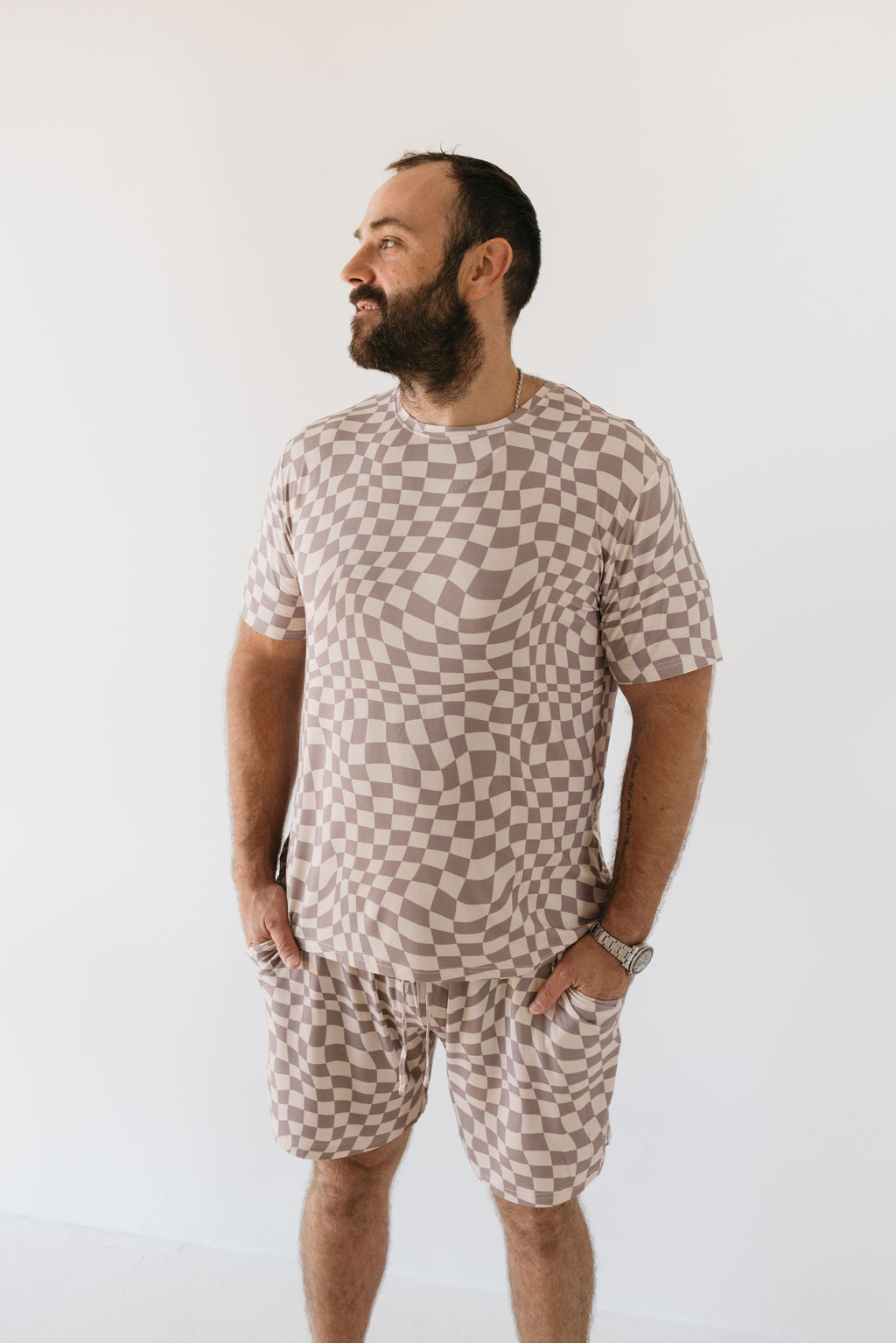 A man with a beard stands against a plain background, wearing the Men's Bamboo Short Pajamas in Smokey Wave by forever french baby. The checkered beige and white attire, crafted from breathable bamboo fabric, features both a t-shirt and shorts. With his hands in his pockets, he gazes to the side, effortlessly combining style and comfort.