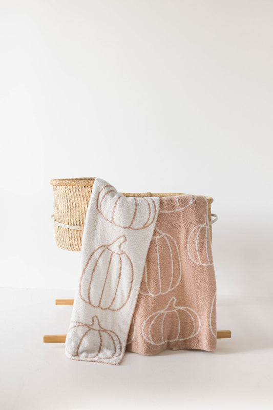 Two cozy, soft blankets from forever french baby's Plush Blanket | Pumpkin Pie collection are draped over a wicker basket. Crafted from microfiber yarn and OEKO Standard 100 certified, the blanket on the left is white with light brown pumpkin outlines, while the one on the right is light brown with white pumpkin outlines, set against a minimalist white background.