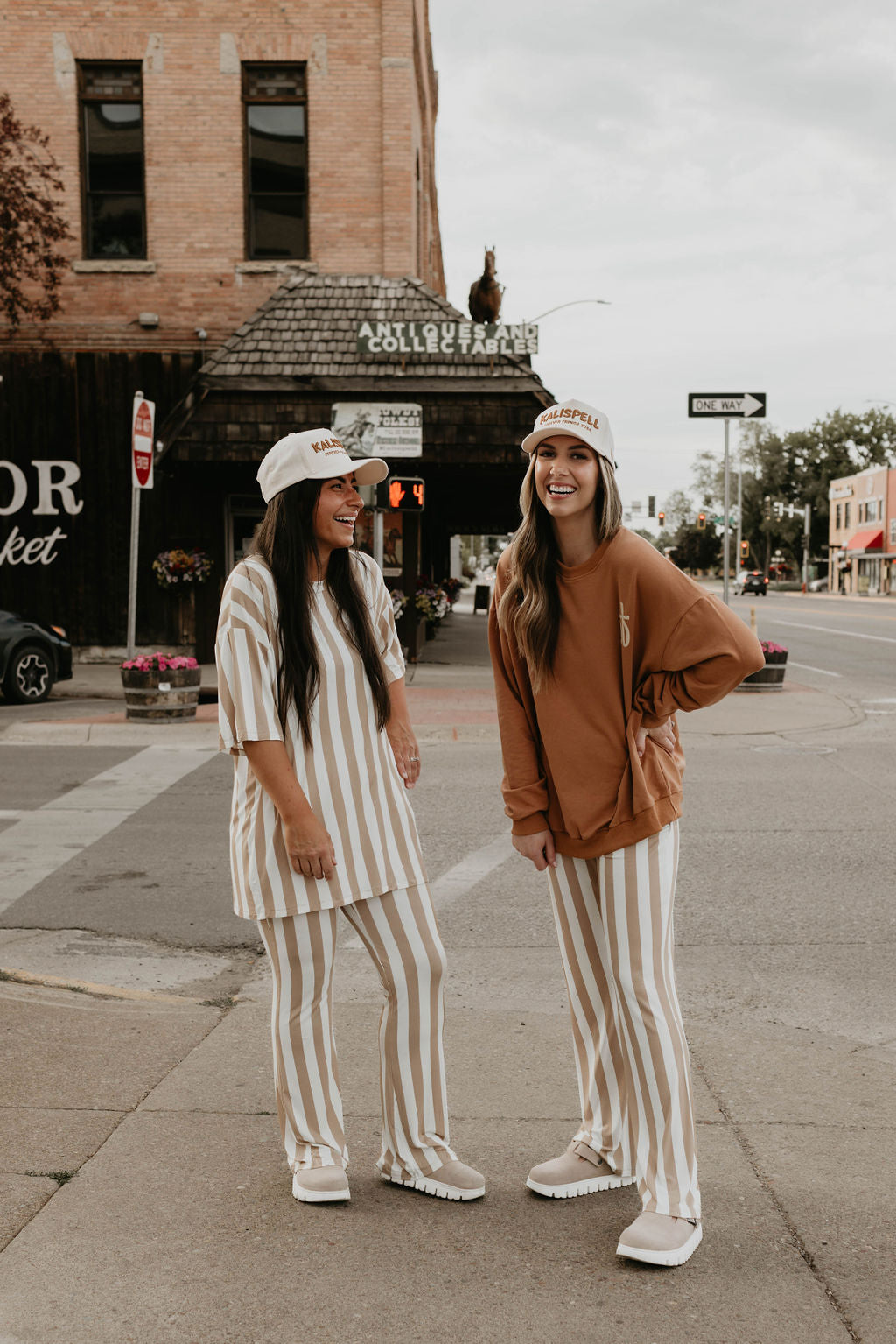 Two women standing on a street corner, both smiling broadly. They are dressed in coordinating outfits featuring striped pants made from 95% cotton and white caps. One wears a striped shirt, while the other sports the "Made It to The Mountains" Adult Sweatshirt by forever French baby. A brick building, street signs, and flowers are visible in the background.