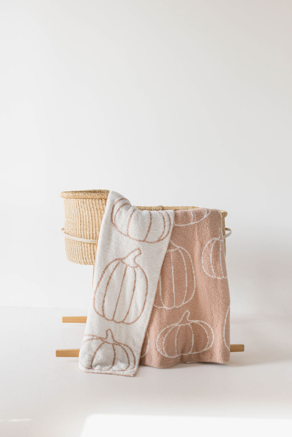 A woven basket holds two soft Plush Blankets from forever french baby, draped over the edge. One blanket is white with an outline of pumpkins, and the other is peach with a similar pumpkin pattern. Made from OEKO Standard 100 microfiber yarn, the scene is set against a white background, creating a simple, clean, and cozy aesthetic.