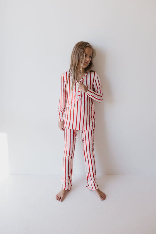 A young girl stands barefoot against a white wall, adorned in the "Pre-Teen Flare Bamboo Pajamas | the Claus" by forever french baby, featuring matching red and white stripes made from breathable bamboo fabric. Her hair falls loosely around her shoulders as she gazes to the side, exuding a relaxed and casual vibe.