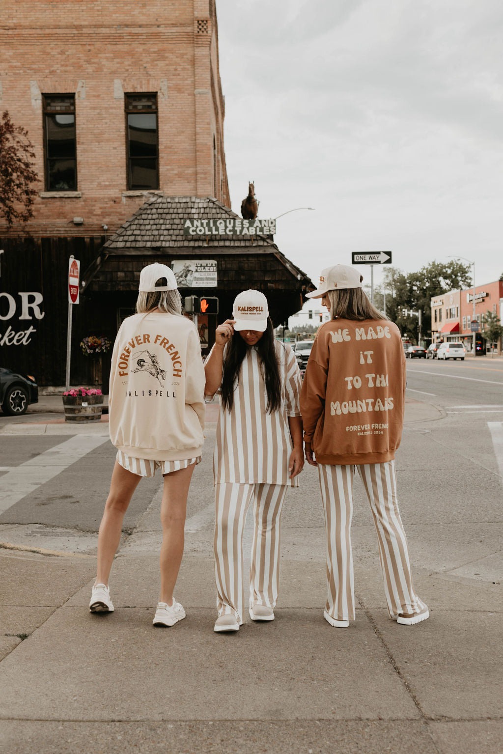 Three people are standing on a street corner in a small town, facing away from the camera. They are casually dressed in matching Kalispell Adult Sweatshirts by forever French baby, made of 95% cotton, and wearing baseball caps. Two buildings and several vehicles, likely for local pickup services, can be seen in the background.