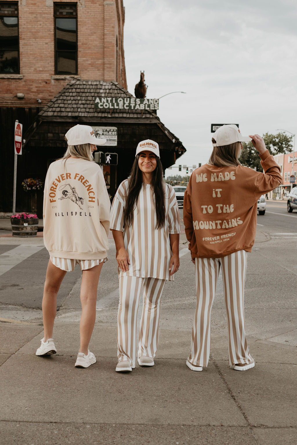 Three women stand on a street, facing away from the camera. They wear matching striped outfits made from 95% cotton and white baseball caps. Text on their clothing reads "FOREVER FRENCH KILISPELL" and "WE MADE IT TO THE MOUNTAINS FOREVER FRENCH." A rustic building is in the background. The sweatshirts they're wearing are from forever French baby, specifically the Adult Sweatshirt called "Made It to The Mountains.