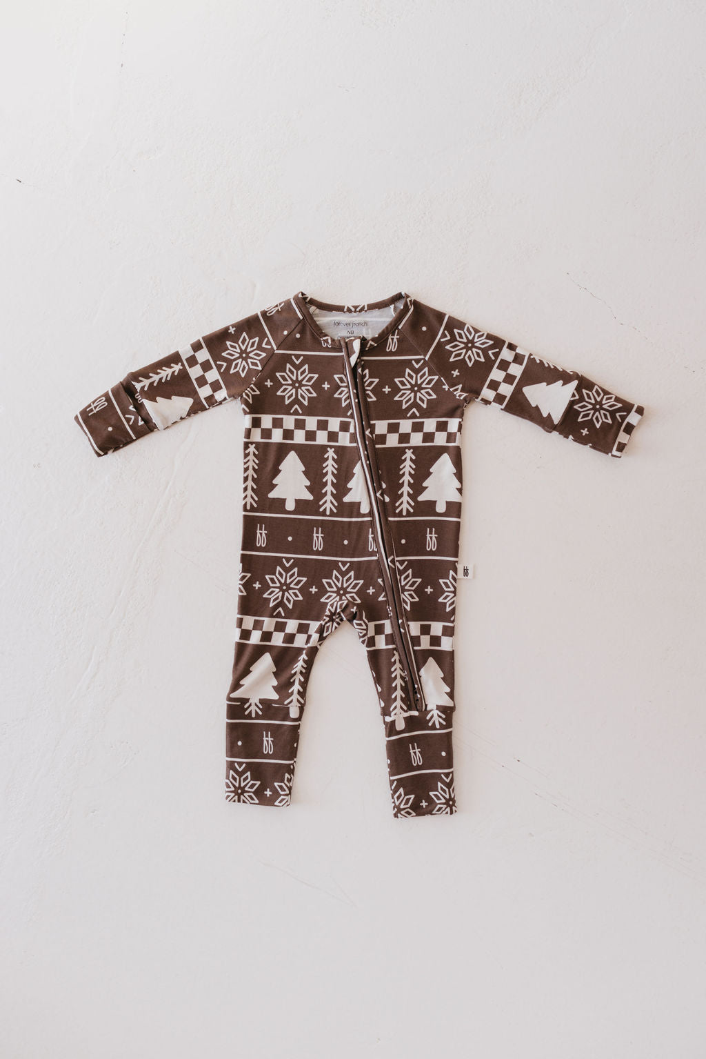 A Bamboo Zip Pajamas from forever french baby, in the Forever Fair Isle design, featuring white holiday-themed patterns like snowflakes, trees, and geometric designs on a brown background, crafted from hypo-allergenic materials and laid flat on a white surface.