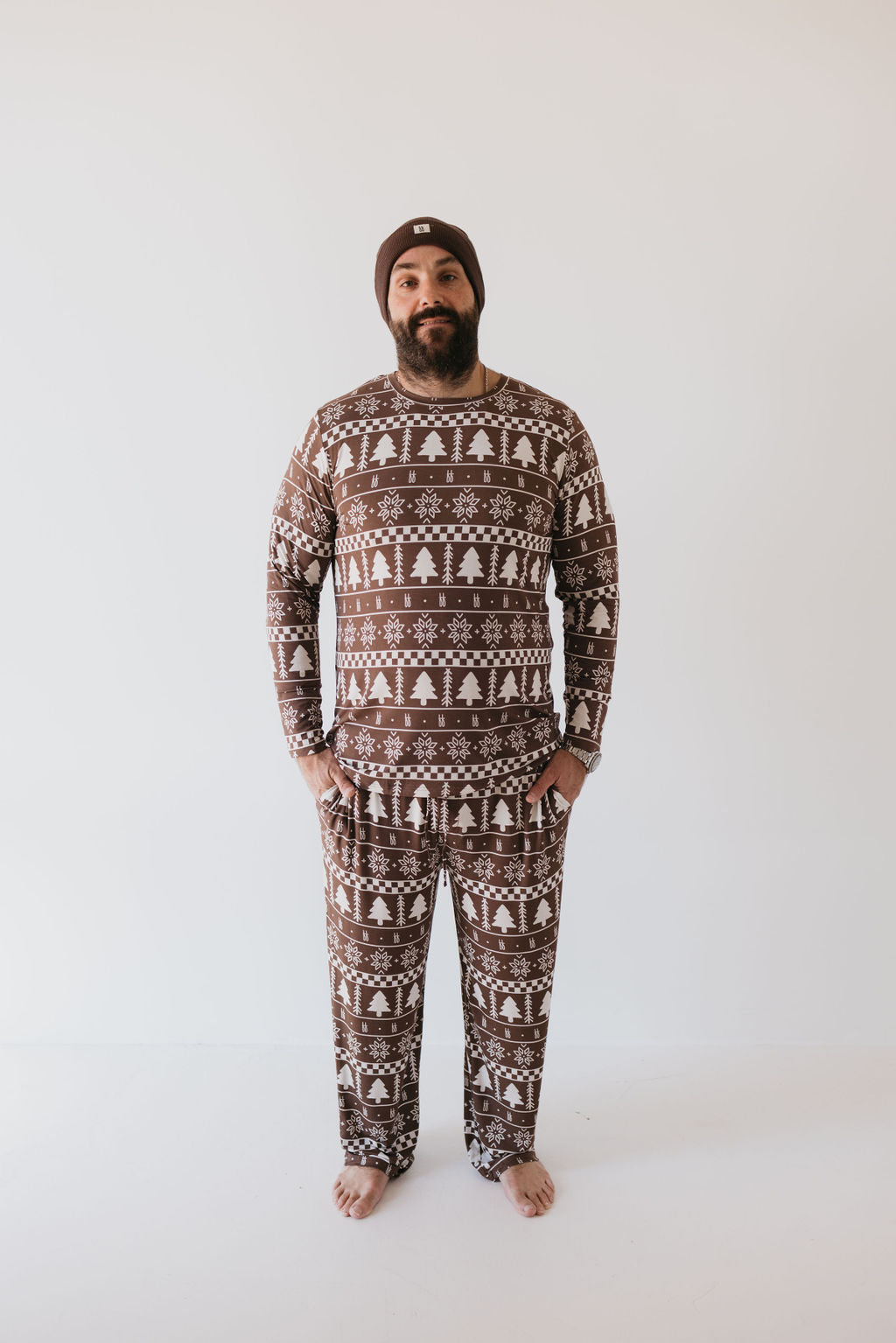 A person stands barefoot against a plain white background, wearing the Men's Bamboo Pajamas by forever french baby, featuring a matching brown winter-themed design with white snowflakes and tree patterns. The cozy attire, crafted from hypo-allergenic bamboo clothing, is paired with a snug brown beanie.