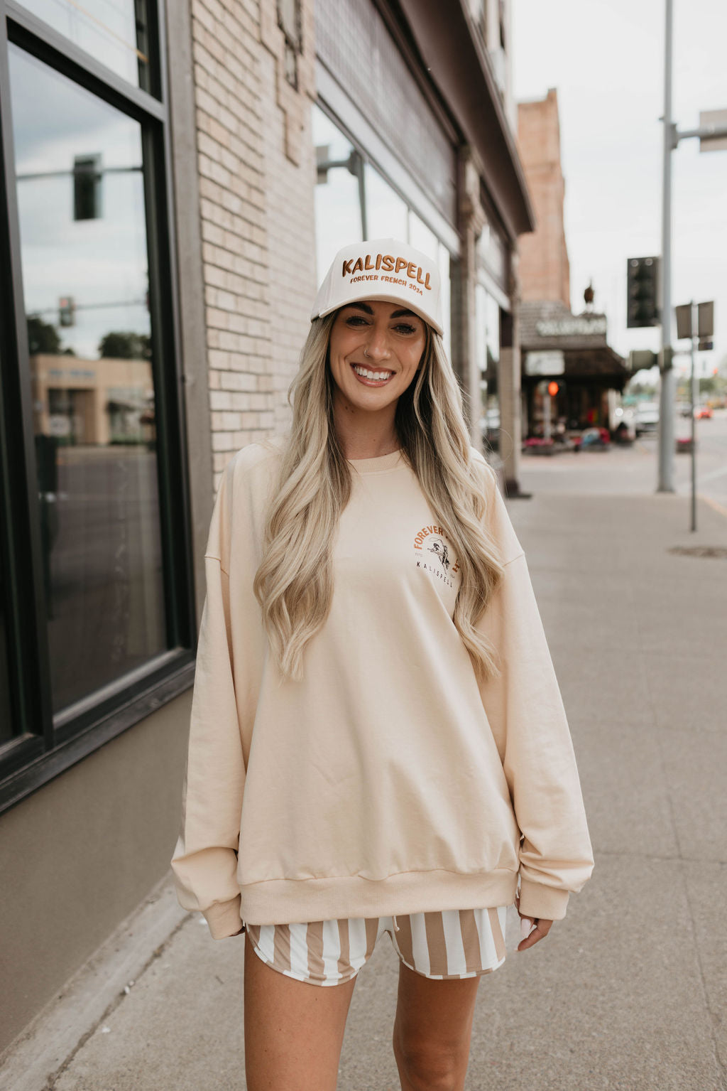 A woman with long blonde hair smiles while standing on a sidewalk in an urban area. She is wearing a beige Adult Sweatshirt by forever French baby, made from 95% cotton and elastane, striped shorts, and a baseball cap that says "Kalispell" on the front. The background includes brick buildings and a street with cars.