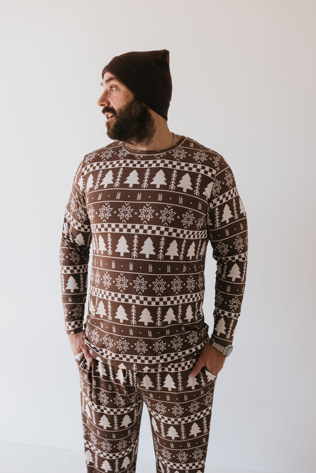 A bearded man is wearing the Forever Fair Isle Men's Bamboo Pajamas from forever french baby, decorated with white snowflakes and tree patterns. His look is completed with a brown beanie as he stands against a plain white background, gazing thoughtfully to the side.