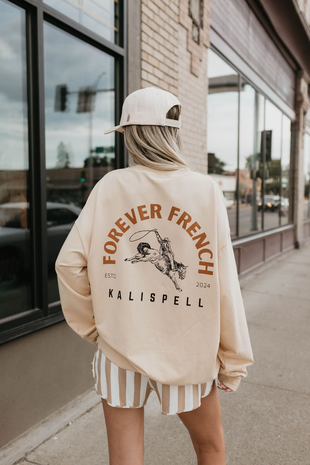 A person wearing a "forever French baby" Adult Sweatshirt | Kalispell in beige with the slogan "Forever French ESTD 2024 Kalispell" and a dog graphic on the back, made of 95% cotton and elastane for enhanced comfort, paired with striped shorts and a cap, strolls along a city sidewalk next to a large window.