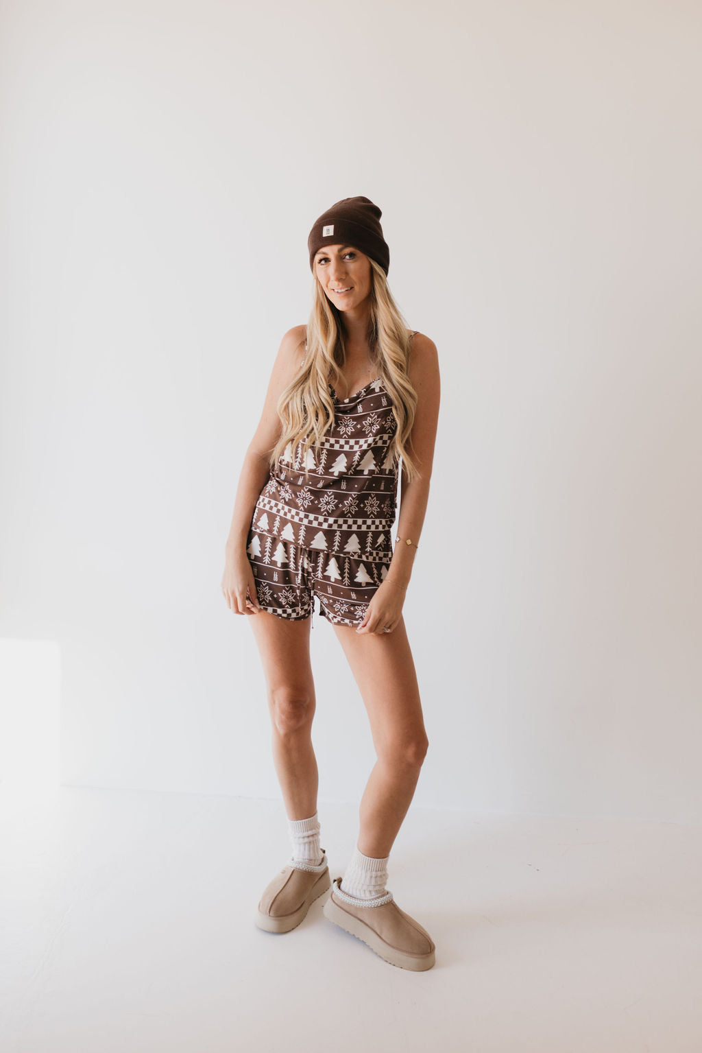 A person wearing the Cami Women's Bamboo Set in Forever Fair Isle by forever french baby stands against a light background. The outfit includes a patterned brown romper made from breathable bamboo fabric paired with a matching brown beanie. They have long hair, a smile, and their hands are resting on their thighs. The look is completed with socks and light-colored shoes.