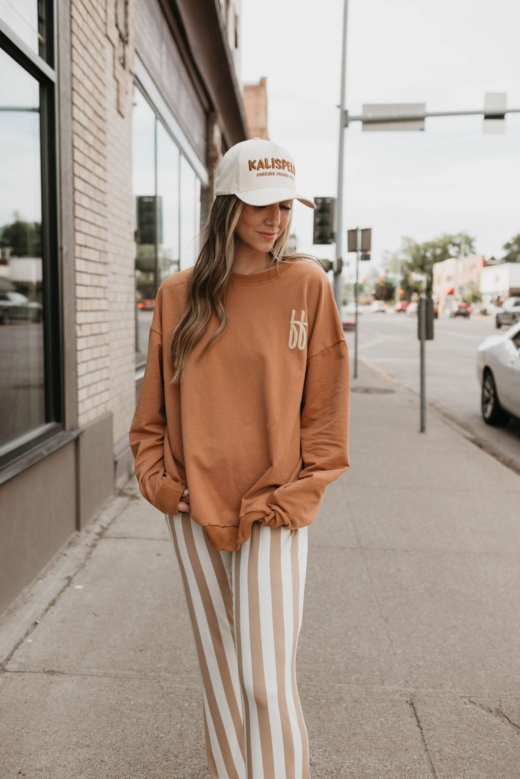 A person with long hair wearing a beige "Trucker Hat | Kalispell" from forever french baby, an oversized brown sweatshirt, and white and beige striped pants stands on a sidewalk beside a brick building. The street next to them, bustling with cars and traffic signals, highlights the convenience of local pickup for nearby stores.