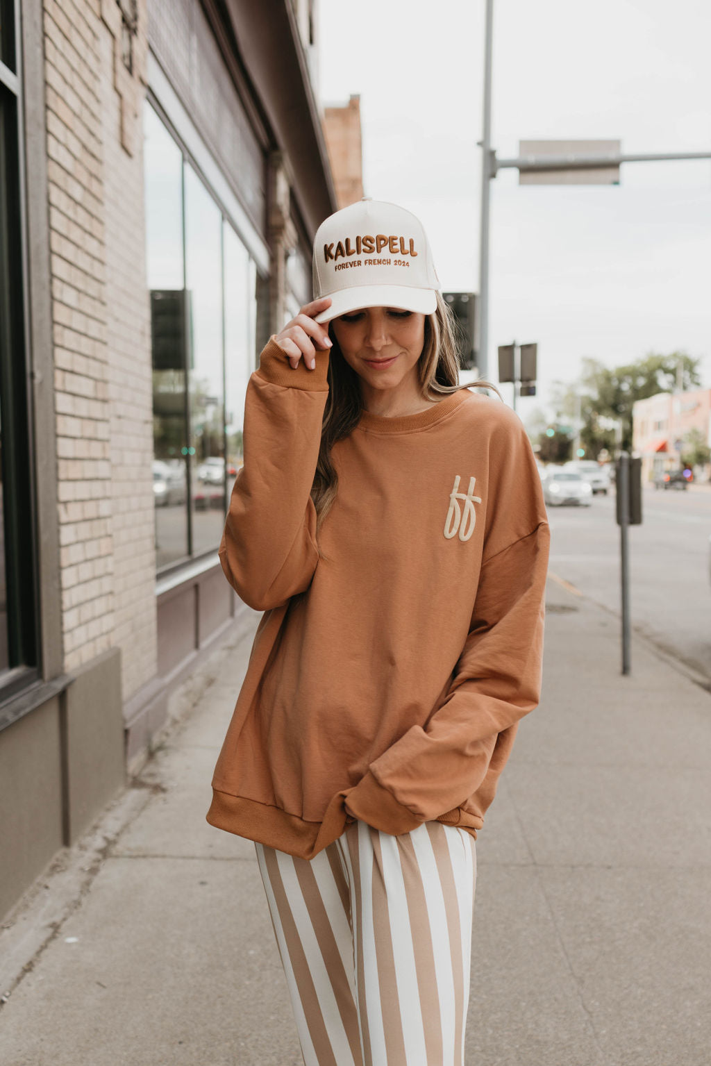 A person wearing a beige "Trucker Hat | Kalispell" by forever french baby and an oversized brown sweatshirt stands on a city sidewalk. They are looking down and holding the front of their hat with one hand. Their brown and white striped pants give off a stylish yet casual vibe, perfect for local pickup or delivery errands.