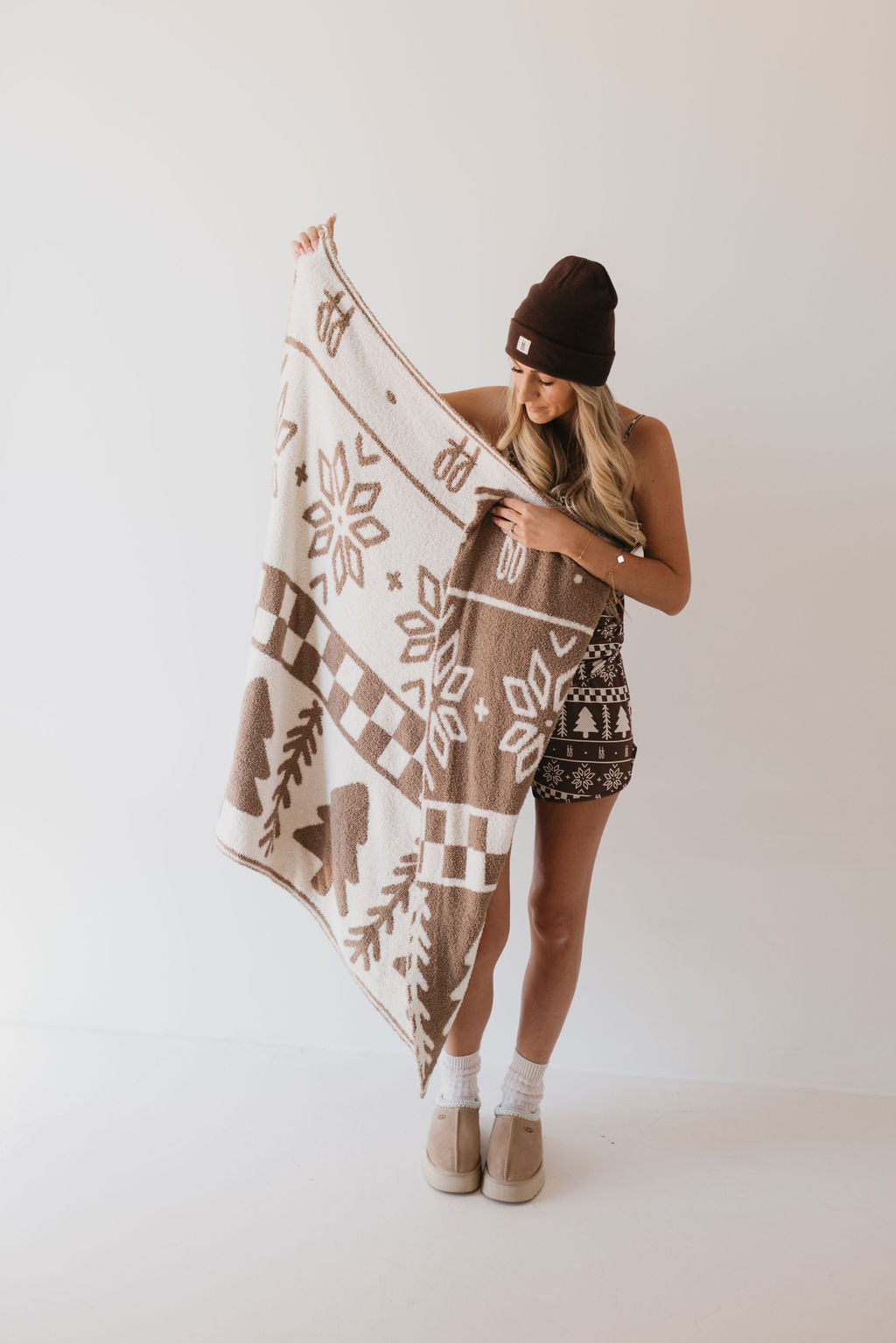 A person wearing a beanie, patterned tank top, and shorts holds an OEKO-TEX certified "Forever Fair Isle" plush blanket by forever french baby, showcasing its brown and white geometric pattern against a plain white background.