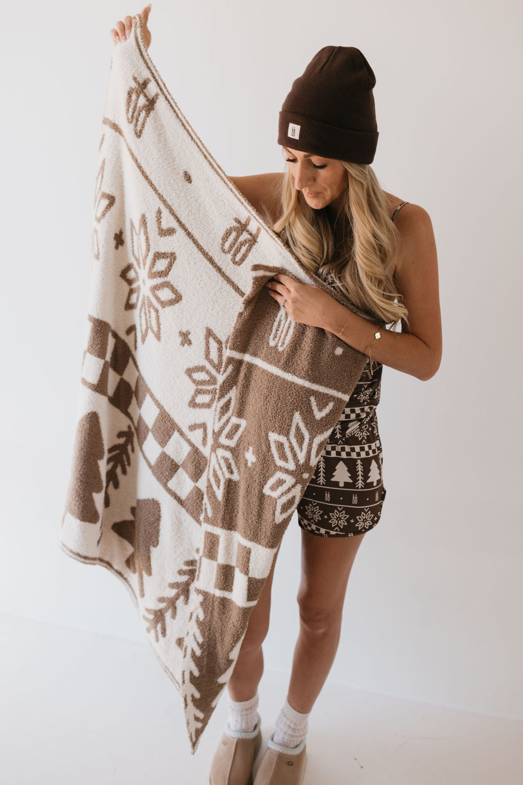 A person wearing a brown beanie and festive outfit smiles while holding the Plush Blanket from the Forever Fair Isle collection by forever french baby, decorated with intricate snowflakes and trees. The scene radiates a cozy, wintery vibe.