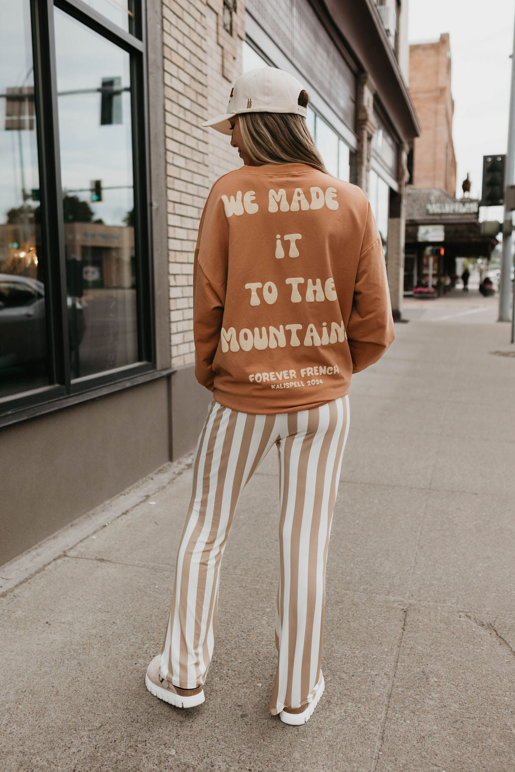 A person wearing a beige cap, the "Adult Sweatshirt | Made It to The Mountains" by forever French baby with white text "WE MADE IT TO THE MOUNTAIN" on the back and "FOREVER FRENCH - CALIFORNIA", paired with white and beige vertically striped pants, walks on a sidewalk near a brick building with large windows.