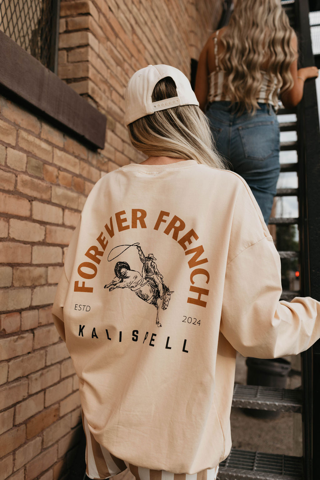 A person with long hair is wearing a light-colored cap and an oversized "Adult Sweatshirt | Kalispell" from forever French baby, which reads "Forever French Kalispell ESTD 2024." The sweatshirt is made of 95% cotton and elastane. They are walking up outdoor stairs behind another individual in jeans and a striped top. The brick wall beside them features a window protected by wire mesh. Local pickup available.