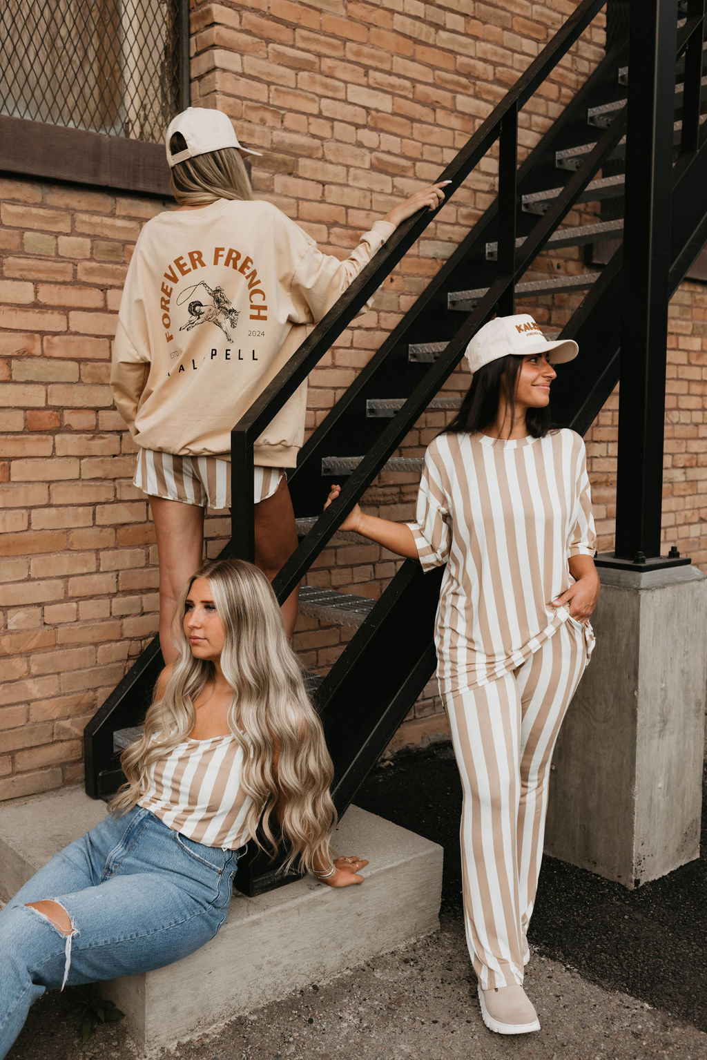 Three women pose near a brick building and staircase. One, in a "forever French baby" jacket made with 95% cotton and elastane, ascends the stairs. Another sits on the step, wearing an Adult Sweatshirt | Kalispell and ripped jeans. The third stands in a striped outfit and cap, leaning against the railing.