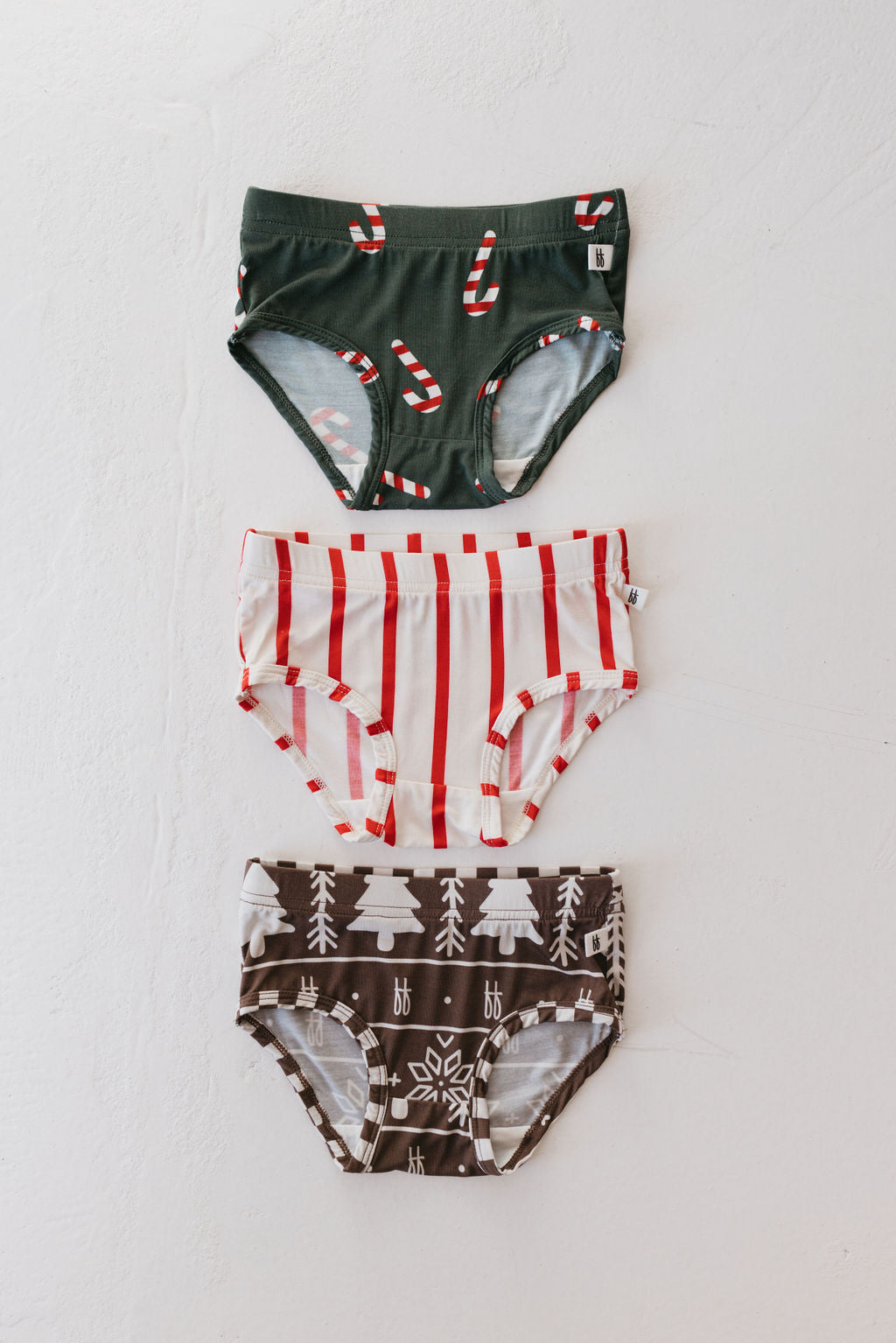 Three pairs of holiday undies from the "Girl's Bamboo Briefs (3 Pack) | 'tis the Season" by forever french baby are laid out vertically on a white surface. The top pair features green fabric adorned with candy canes, the middle pair shows off a white base with red stripes, and the bottom pair boasts brown Christmas trees and snowflakes—the perfect choice for children's briefs during festive celebrations.