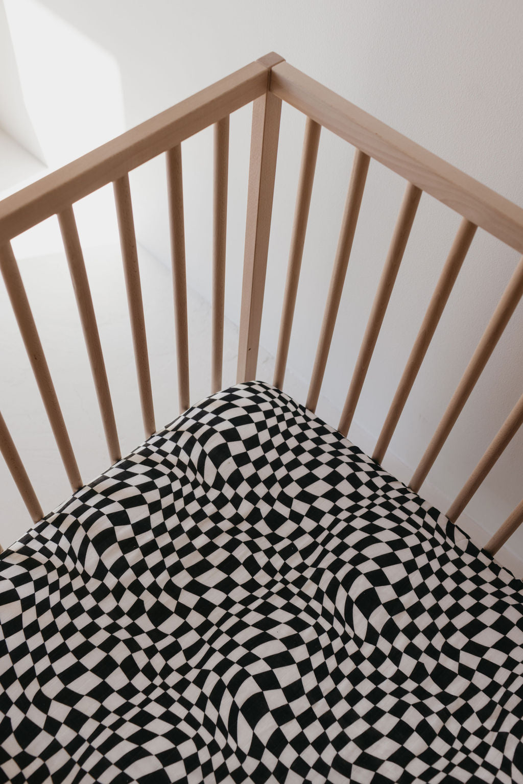 An overhead view showcases a wooden crib with vertical slats, adorned with the Muslin Crib Sheet in Black & White Wavy Checkerboard by forever french baby. The sheet's pattern creates an optical illusion effect with wavy distortions on the crib mattress. A plain white background wall enhances the serene nursery ambiance.