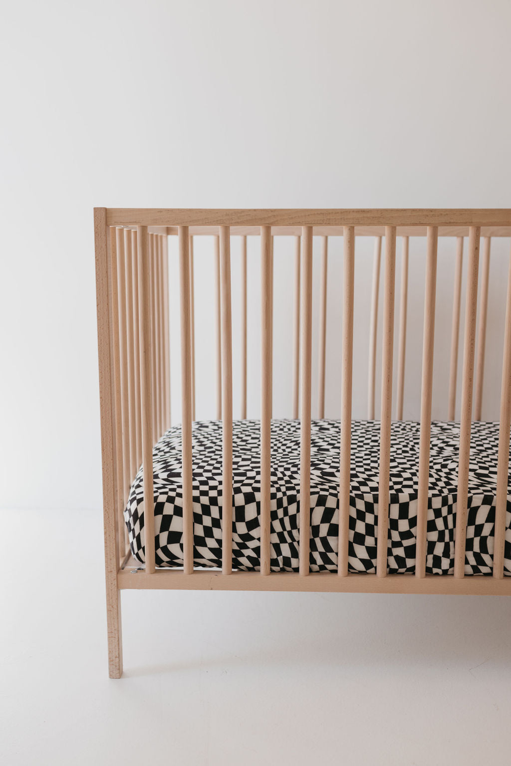 A wooden crib stands on a white floor with a minimalist design. The crib mattress, adorned with the Muslin Crib Sheet in Black & White Wavy Checkerboard by forever french baby, adds contrast to the light-colored wood and surrounding space, making it a charming focal point in the nursery.