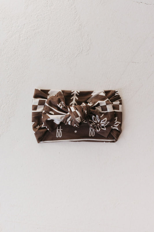 A neatly folded Forever Fair Isle Bamboo Head Wrap by forever french baby, showcasing white geometric and tree patterns on a brown fabric with a large bow in the center, rests on a light textured surface. This adjustable head wrap is perfect as a stylish and comfortable accessory for babies and toddlers.