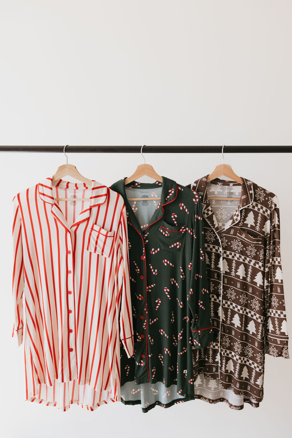 Three festive, eco-conscious shirts on display: one with red and white stripes, another green adorned with candy canes, and a final brown featuring white Christmas trees and snowflakes, all hanging against a plain white backdrop.