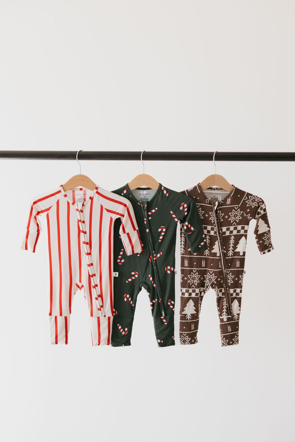 Three *Bamboo Zip Pajamas | the Claus* from *forever french baby* hang on a clothing rod. These breathable baby pajamas showcase diverse patterns: red and white stripes, green adorned with candy canes, and brown decorated with Christmas trees and snowflakes.