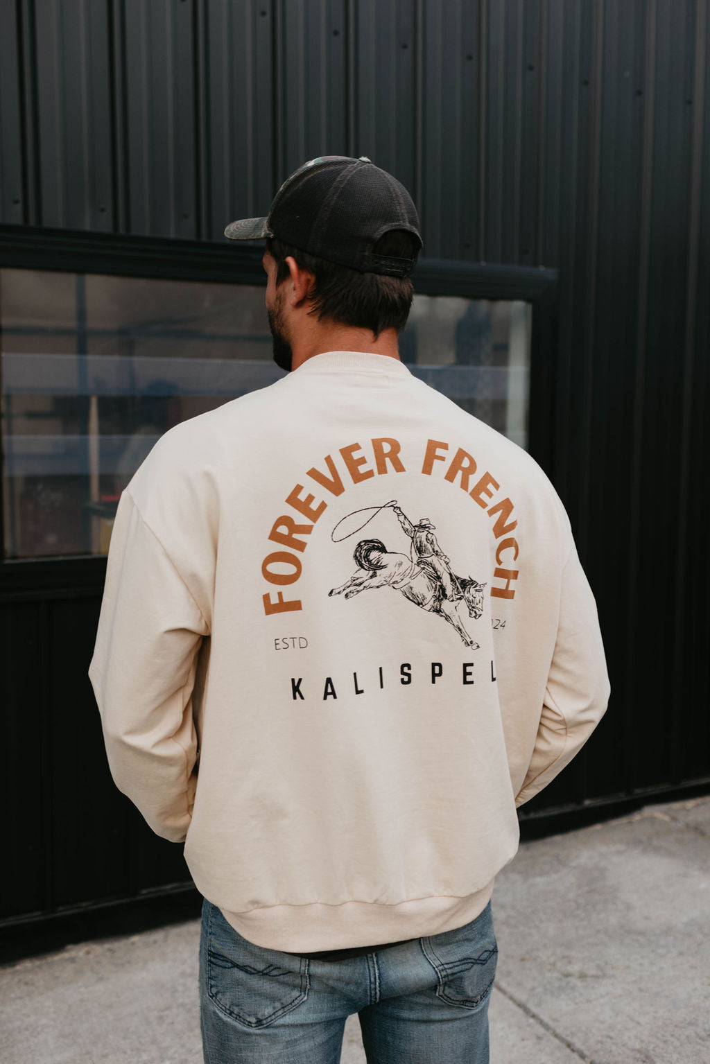 A man stands with his back to the camera, wearing a black cap and a beige sweatshirt. The back of the 95% cotton sweatshirt from forever French baby features "FOREVER FRENCH" above a graphic of a person climbing a mountain, with "ESTD KALISPELL" and a small inscription beneath. Local pickup available.