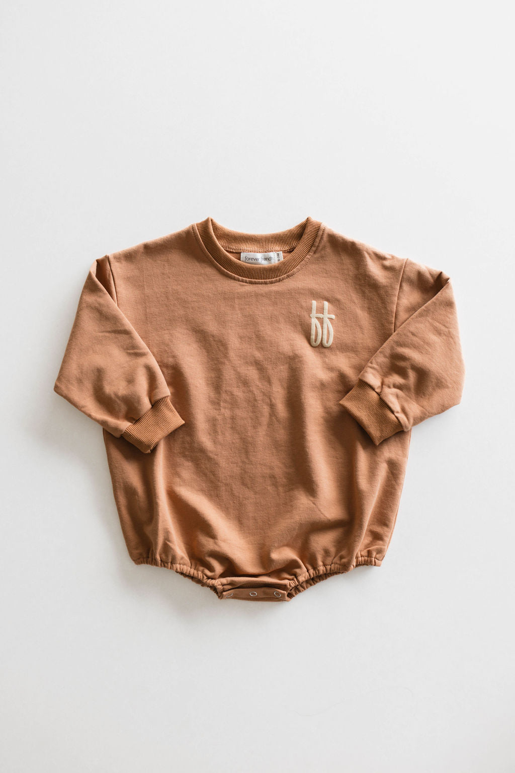 Introducing the "Made It to The Mountains" Baby Romper by forever French baby, crafted from light brown cotton fabric with an embroidered "bb" logo on the chest. This long-sleeve romper features ribbed cuffs, a snap button closure at the bottom, and a simple crew neckline, all showcased against a plain white background.