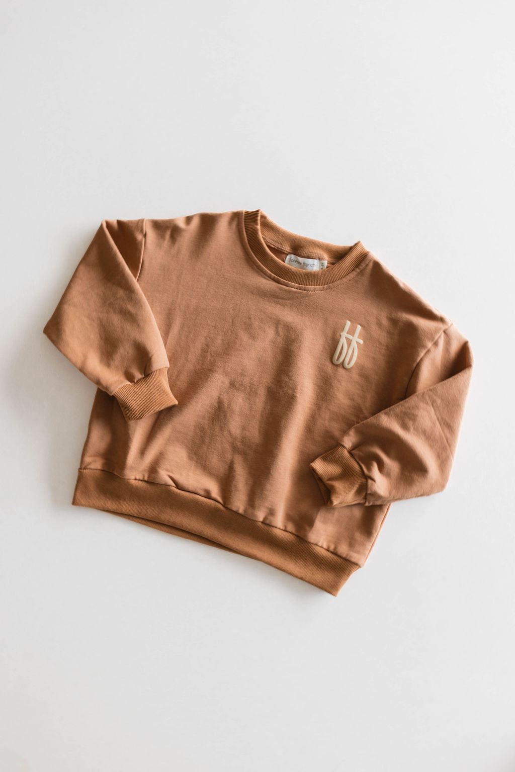 A light brown, long-sleeved crew neck sweatshirt from forever French baby's "Children's Sweatshirt | Made It to The Mountains" collection is laid flat on a plain white background. Made from 95% cotton with a touch of elastane, this relaxed-fit sweatshirt features a small, white "ff" logo embroidered on the left chest. Available for local pickup.