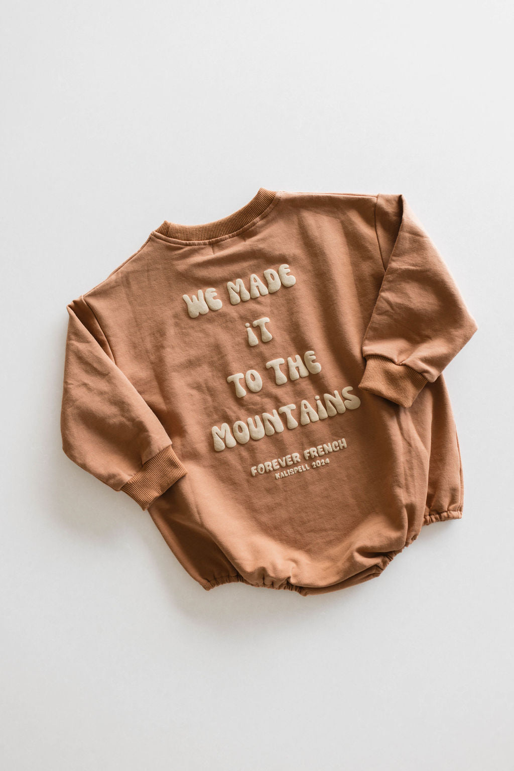 The Baby Romper from forever French baby, made from a soft cotton blend with a touch of elastane for added stretch, features puffy white text on the back that reads "WE MADE IT TO THE MOUNTAINS" and "FOREVER FRENCH, EXPIRY DATE UNKNOWN." This brown long-sleeved romper is displayed laid flat on a light gray background.